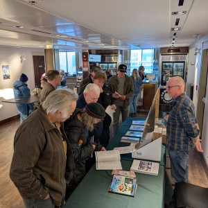 In the foreground, a small crowd of people are gathered in the Market Hall Performing Arts Centre lobby. Visuall artist Rob Niezen is seen wearing a plue and yellow plaid shirt and glasses. He has his hands clasped behind his back and is standing behind a table covered by a dark green tablecloth filled with prints. Several people dressed in coats and hats are gathered around the table. One person in a black coat and green hat looks through a pamphlet or small book on the table, she is standing slightly behind a man in a brown jacket. There is a bar in the background of the picture with a staff member serving two indivisudals. On the left wall a "Washroom" sign is visible as well as a door. The room is well light, with windows and overhead lighting and the atmosphere appears to be calm and welcoming.