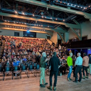 An audience gather at Market Hall Performing Arts Centre ahead of a film screening, the space is well lit with a combination of cool and warm lights. There four rows of chairs on the floor, as well as auditorium and balcony seating. Most individuals are already seated while some make their way to their seats, and others engage in conversation. A tech booth is visible in the left upper corner with two people are partially visible. The venue features exposed beams, and high ceilings, creating an intimate yet spacious setting.