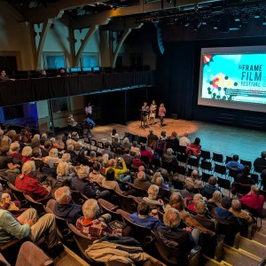 A large audience is seated in the Market Hall Performing Arts Centre attentively listening to three presenters on stage at the ReFrame Film Festival. Lit by a spotlight, one presenter stands slightly in front of the others speaking at the podium. All three presenters are in front of a large screen displaying the festival's branding, including a geometric floral design and text. Captions for accessibility are visible on the screen. The image is shot from behind the audience, with only the backs of those seated visible. In the wings the the left of the presenters a person using a mobility aid is visible as well as a volunteer in a pink shirt. Three individuals are visible seated on the left side of the balcony watching the presenters.