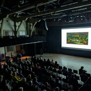 A large audience is seated in the Market Hall Performing Arts Centre, attentively watching a film screening at ReFrame Film Festival. The audience is dimly lit, with the screen as the primary light source. On screen is a rectangular image made up of green, yellow and brown bordered by white. The venue features exposed beams, high ceilings, and tiered seating, creating an intimate yet spacious setting