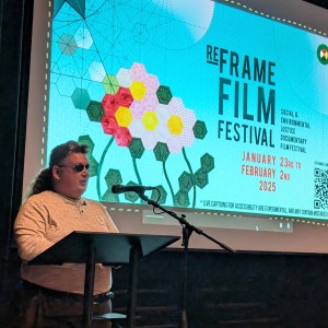 Jason King, Marketing and Programming Coordinator Council for Persons with Disabilities (CPD),is wearing a white longsleave shirt, grey pants, and sunglasses while speaking ahead of a film screening. Jason is standing in front of a podium and a mic stand, behind them a screen displays the ReFrame Film Festival branding, including geometric floral artwork, event dates, and a QR code. Jason has a hair is short at the front with a long ponytail in the back.