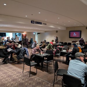 Numerous individuals sit at tables in the Cogeco Lounge located in Showplace. The tables are black with black chairs and people engage in conversations with those seated with them. On the left there is a bar serving snacks and drinks, there are several people waiting in line. The atmosphere is casual and friendly.