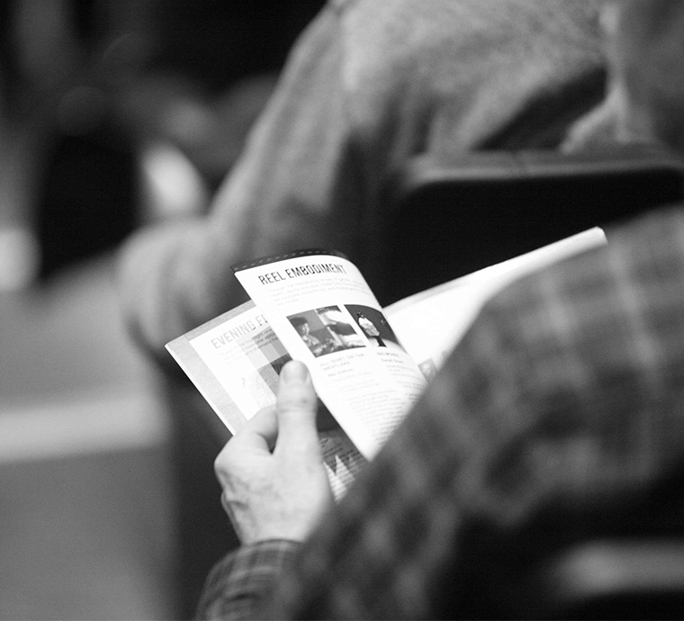 A square black and white image taken over the sholder of a person in a plaid shirt flipping through the ReFrame 2025 program in a theatre seat with another person in front of them.