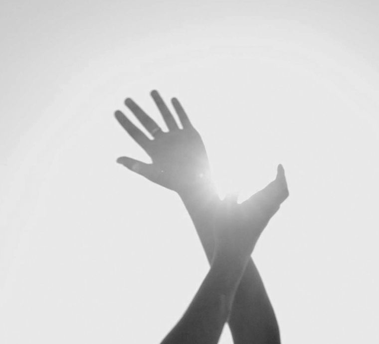 A black and grey image of two hands crossed and reaching into a brightly lit sky. 