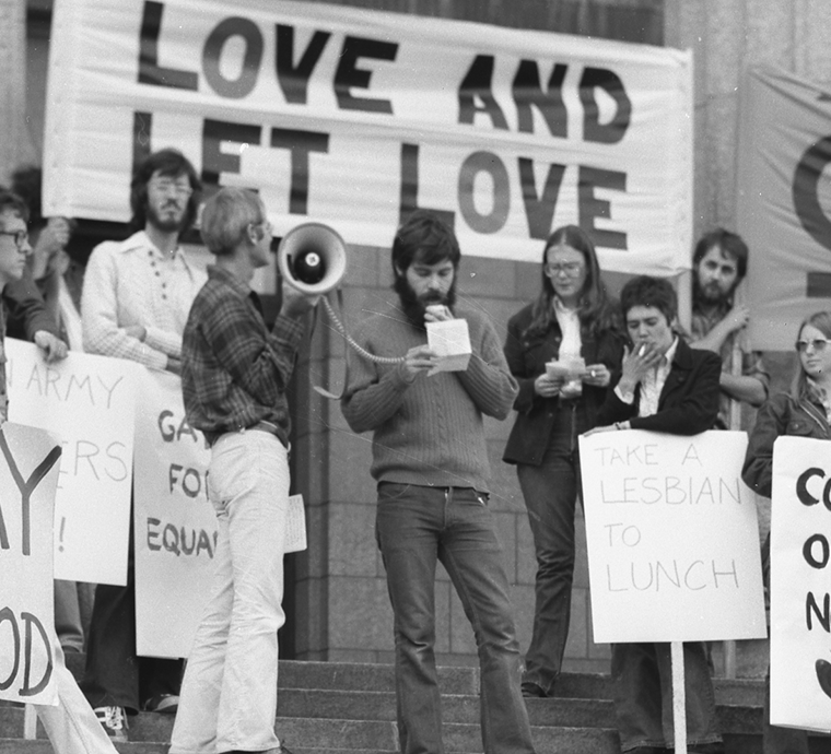 A group of people stand on the steps of a building during a protest or rally. They hold various signs, including one large banner in the background that reads “LOVE AND LET LOVE.” Other visible signs say “GAYS FOR EQUALITY” and “TAKE A LESBIAN TO LUNCH.” One person in a plaid shirt holds a megaphone, while another person beside him reads from a notepad into a microphone. Most individuals appear to be young adults, dressed in casual 1970s-style clothing. The atmosphere suggests an LGBTQ+ rights demonstration.