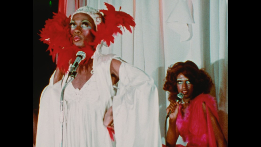Two performers are on stage in front of white curtains. The person in the foreground wears a dramatic white gown with flowing sleeves, a headpiece adorned with white lace and large red feathers, and bold makeup with red lipstick and blue eyeshadow. They are singing or speaking into a microphone, hand on hip, looking upward. The second performer in the background is dressed in a vibrant pink, semi-sheer outfit with ruffled details, also holding a microphone. Their makeup includes bright eyeshadow and lipstick, and they have voluminous auburn hair. The setting suggests a stage performance, likely in a cabaret or drag show.