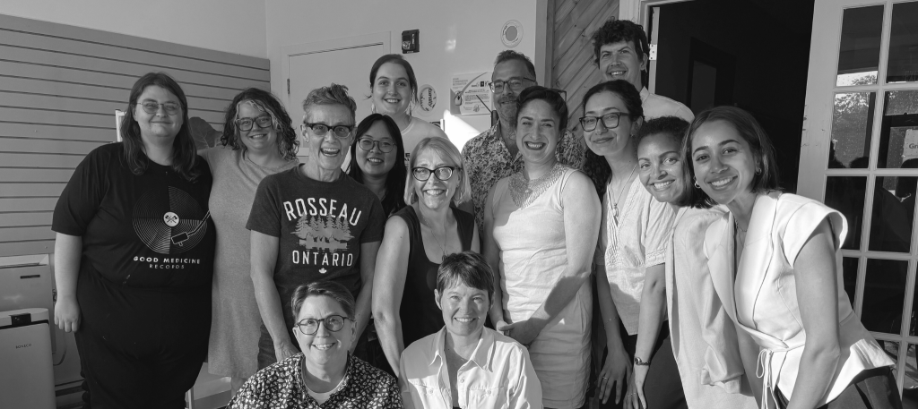 A group of fifteen people pose closely together indoors, smiling at the camera. They are standing and crouching in two rows, forming a tight, friendly group. The individuals are of varied ages, hairstyles, and attire. Some wear glasses, and clothing ranges from casual t-shirts to collared shirts and blouses. The lighting suggests natural sunlight coming from the right side of the image, casting shadows and highlights across their faces and clothing. Behind them is a white wall with horizontal paneling, a door, and windows with visible reflections.