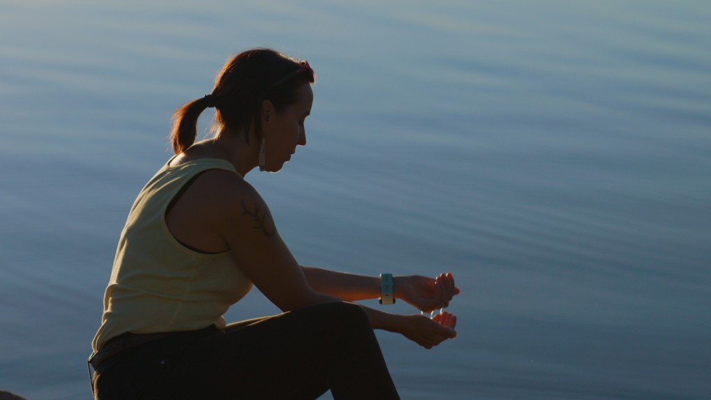 A young person with a pony tail sits beside a large still body of water. They are in profile and almost silhouetted with their hands cupped in front of them. 