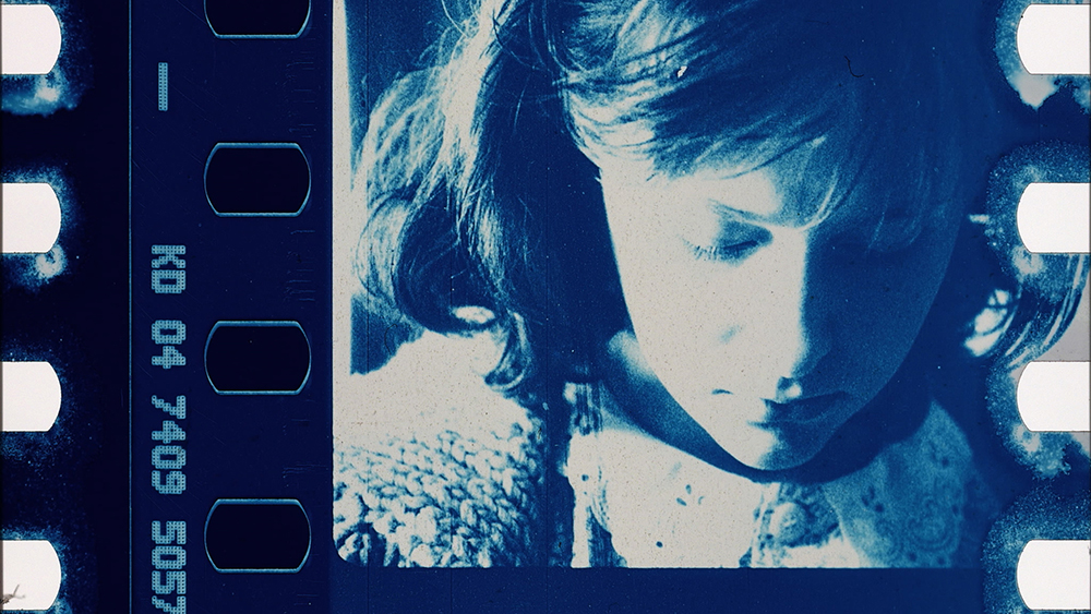 A cyanotype film still of a young girl's face looking down from Kelly Egan's quilt work. Sproket holes and film code are visable at the edges.