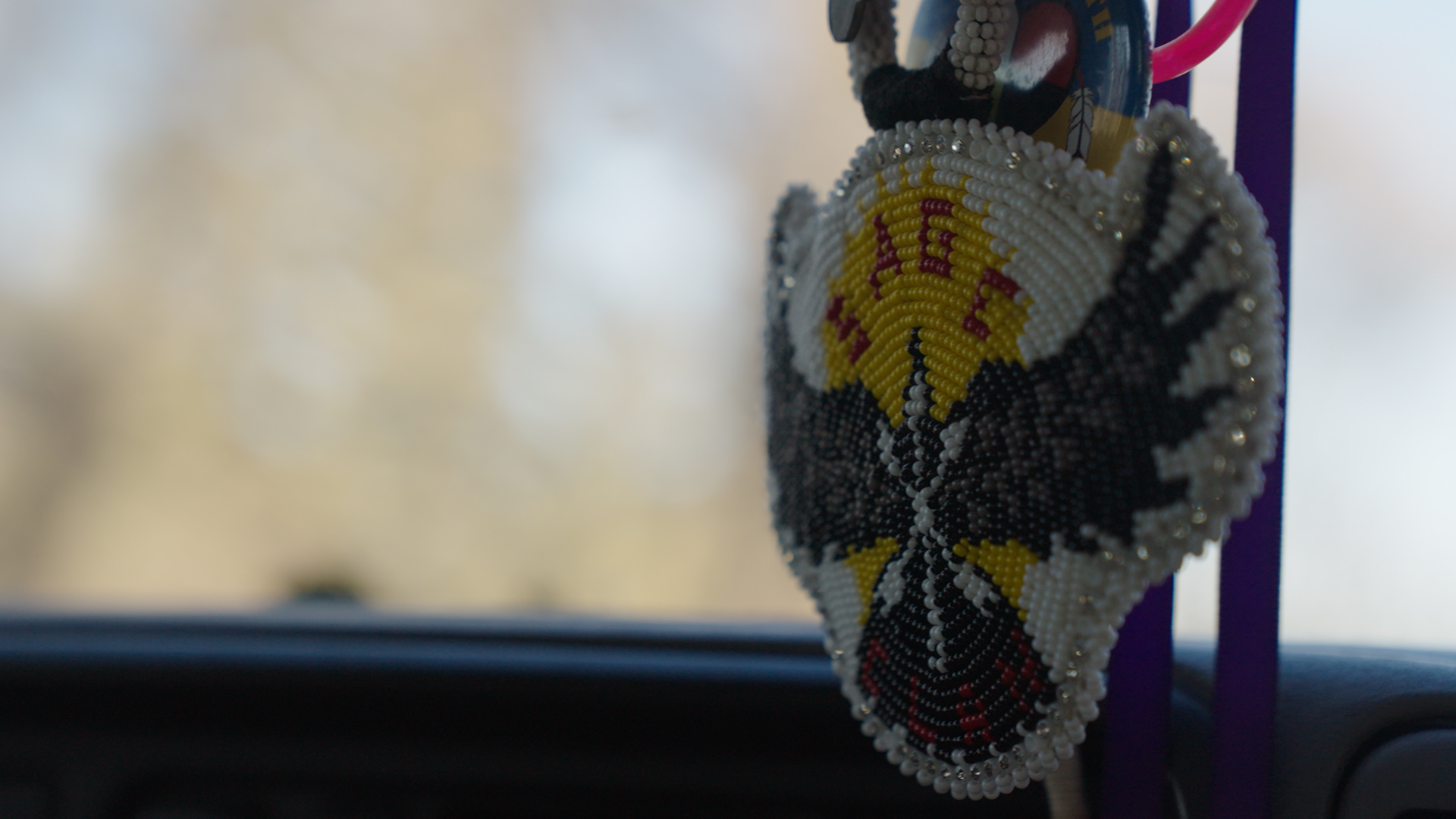 Image from micro credential film #skoden. A picture taken inside a car looking out the winsheild at blurry scenery. Focus is on beadwork, apparently hanging from the rearview mirror, depicting an eagle with yellow and white background and the word SAGE in red.