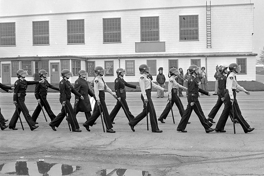 Black-and-white photograph of a line of uniformed police officers wearing helmets and carrying batons, marching in formation during the Fleck Strike in Canada, with an industrial building and onlookers in the background.