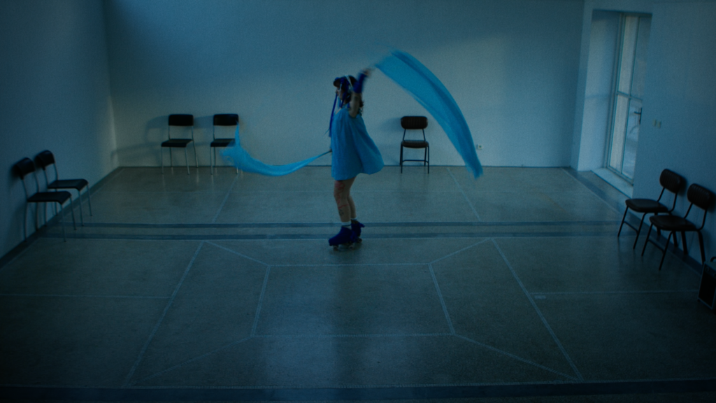A person on roller skates twirling blue fabric in a dimly lit room with opne wooden floows and a few shairs against the walls.
