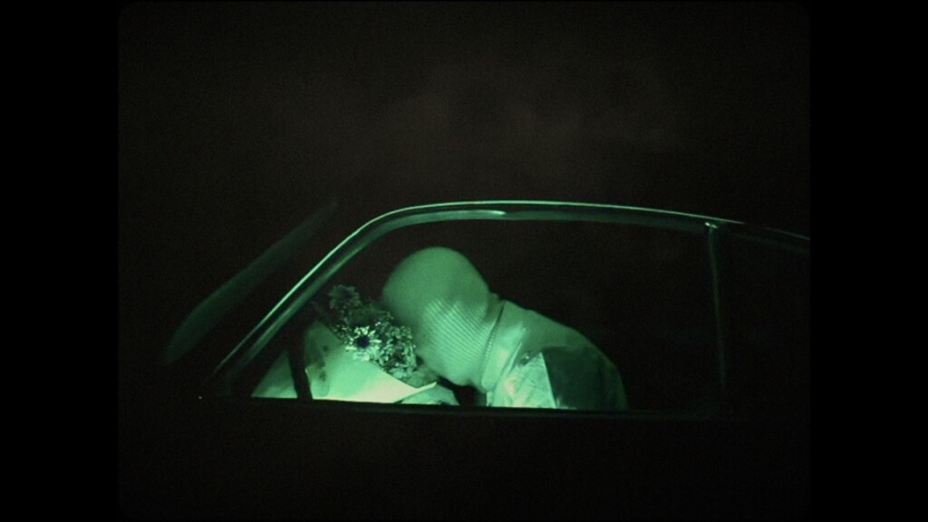 A person wearing a white balaclava is illuminated in green light inside the drivers seat of a car. Their face is pressed into a bouquet of flowers resting against the steering wheel.