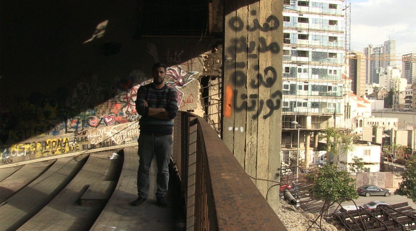A person stands on a balcony with their arms crossed, surrounded by graffiti-covered walls. Behind them, a cityscape with various buildings in different states of construction and maintenance is visible. The contrast between the neglected foreground and the bustling city background highlights a narrative of urban resilience and perhaps social issues. The sunlight casts shadows, adding to the gritty ambiance of the setting.