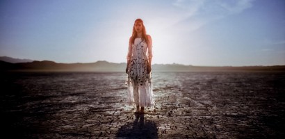 A person stands at the center of a vast, cracked desert landscape under a bright sky with the sun directly behind, casting a long shadow forward. They wear a mid-length dress with a floral pattern and appear barefoot. The horizon is lined with mountains, and the lighting suggests either dawn or dusk due to the warm tones and long shadows. The ground is parched and textured, highlighting the dry environment.