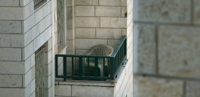 A lone white plastic chair sits on a small, narrow balcony with green railings. The balcony is part of a building with a facade of large, beige stone tiles. The viewpoint is from an adjacent building, capturing the scene at a slight angle, which gives a voyeuristic glimpse into this quiet urban space.
