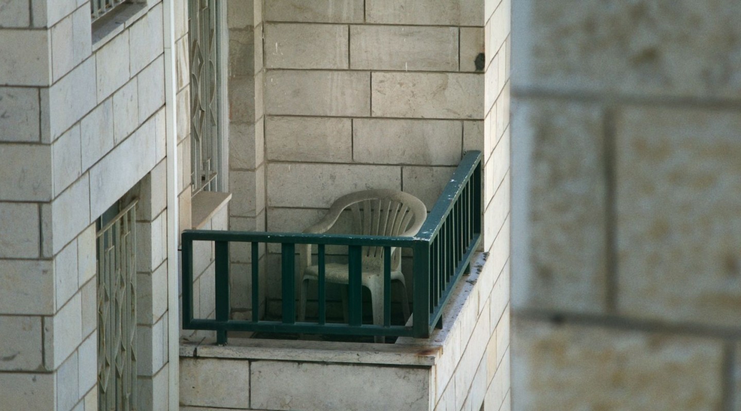 A lone white plastic chair sits on a small, narrow balcony with green railings. The balcony is part of a building with a facade of large, beige stone tiles. The viewpoint is from an adjacent building, capturing the scene at a slight angle, which gives a voyeuristic glimpse into this quiet urban space.