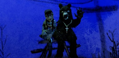 A person wearing a cap, glasses, and a cross necklace is seated beside a large figure in a bear costume. The bear figure is standing on two legs, with one arm raised and the other extended forward, displaying claws. Both figures are set against a blue-tinted backdrop with silhouetted foliage, creating a surreal, nocturnal scene.