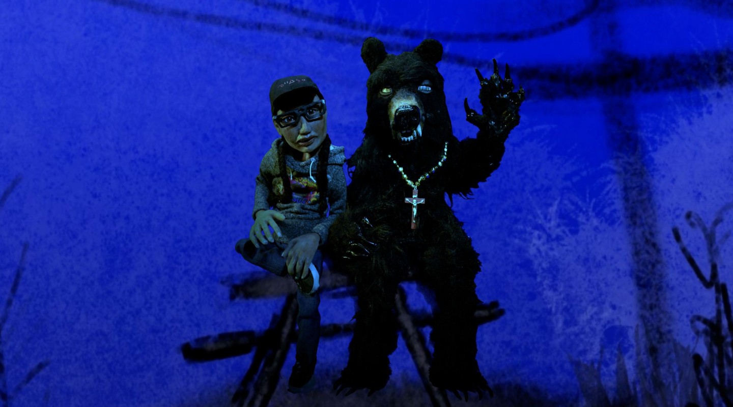 A person wearing a cap, glasses, and a cross necklace is seated beside a large figure in a bear costume. The bear figure is standing on two legs, with one arm raised and the other extended forward, displaying claws. Both figures are set against a blue-tinted backdrop with silhouetted foliage, creating a surreal, nocturnal scene.