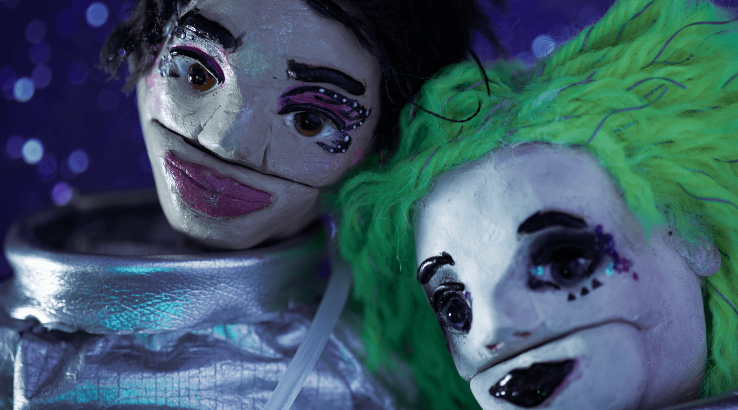 Two puppet-like figures with painted faces and exaggerated makeup are positioned close together against a bokeh background with purple and blue hues. The figure on the left has black hair and pink makeup, while the one on the right has bright green hair and darker makeup around the eyes. The facial expressions are fixed and stylized, with a handmade quality to the crafting of their features.