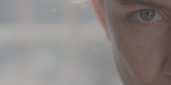 Close-up of the left side of a person's face, focusing on a clear blue eye. The image captures detailed textures such as skin and eyelashes, with the rest of the face softly out of focus. The background is indistinct, providing a neutral setting that highlights the eye.