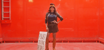 A person stands confidently in a power stance against a vibrant red background. They are dressed in a black leather jacket over a t-shirt with the phrase "Ignorance is Your Enemy" written on it, a short brown skirt, and thigh-high tan boots. Their right hand is on their hip and they wear a black beret with a gold emblem. Beside them is a placard leaning against their leg with "POWER TO THE PEOPLE" in bold letters. The environment suggests an urban setting, possibly a staged or artistic setup given the uniform red colour and lighting.