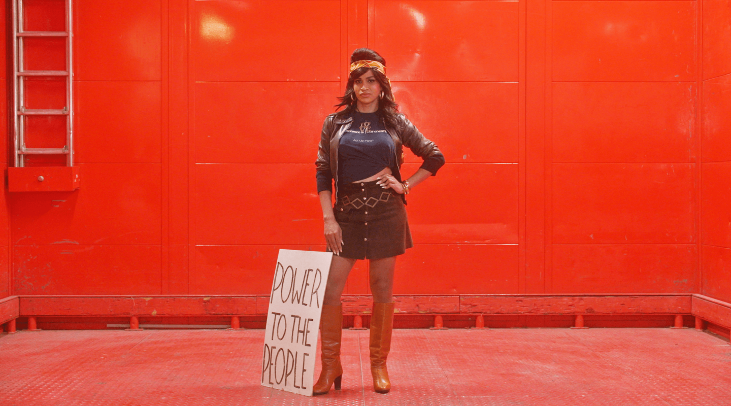 A person stands confidently in a power stance against a vibrant red background. They are dressed in a black leather jacket over a t-shirt with the phrase "Ignorance is Your Enemy" written on it, a short brown skirt, and thigh-high tan boots. Their right hand is on their hip and they wear a black beret with a gold emblem. Beside them is a placard leaning against their leg with "POWER TO THE PEOPLE" in bold letters. The environment suggests an urban setting, possibly a staged or artistic setup given the uniform red colour and lighting.