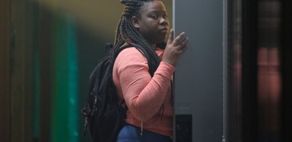 A person with braided hair is standing at a locker, looking over their shoulder with a contemplative expression. They are wearing casual clothing and a backpack, indicating they could be a student. The hallway in the background is blurred.