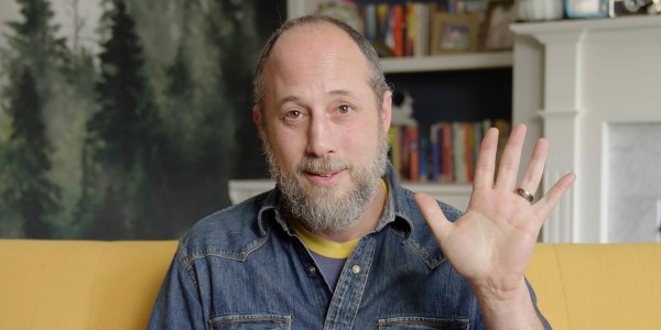 An individual with a beard and a receding hairline is sitting on a yellow couch, wearing a denim jacket over a yellow shirt. They are raising their right hand with an open palm towards the camera in a greeting or waving gesture. The person is smiling and looking directly at the camera. In the background, there's a bookshelf filled with books and a painting of a forest scene, suggesting a cozy, lived-in space. The lighting is bright and even, indicative of an indoor setting.