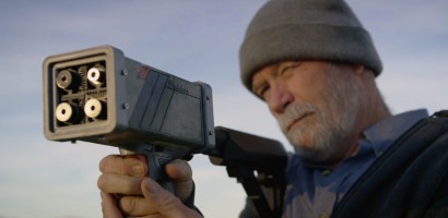 An individual, likely a mature adult given the visible gray beard and wrinkles, is aiming a laser gun. The device is held up to their face, and their focused gaze is directed towards the target. They are wearing a gray beanie and a dark jacket, and the background is softly blurred with a clear sky, indicating an outdoor setting during the day. The device’s detailed mechanics are visible, emphasizing a the advanced technology.
