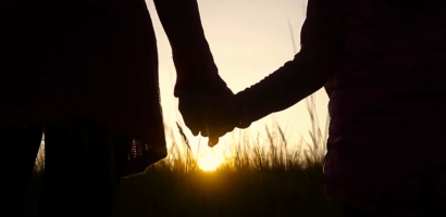 Two individuals are holding hands against a sunset backdrop with a field of tall grasses. The sun is low on the horizon, silhouetting the figures and creating a warm glow that outlines their joined hands. The image captures a moment of connection or partnership in a serene, natural setting.