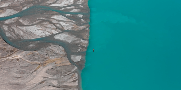 An aerial image that captures a striking natural contrast between a turquoise blue body of water and a greyish, textured landscape possibly of sand or silt. In the water, a small figure in a kayak, provides a sense of scale, emphasizing the vastness of the surroundings. The water's vibrant hue suggests it may be rich in minerals or glacially fed, while the sinuous patterns in the grey area indicate the movement of water over time. The scene is serene and appears remote.