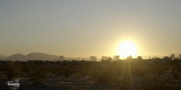 A sunset over a desert cityscape with the sun low in the sky, casting a warm glow and creating a silhouette of the mountains in the background. There are outlines of buildings and palm trees against the light, and the foreground shows arid terrain with sparse vegetation typical of a desert. In the lower left corner, there's text that reads "HOW TO POWER A CITY," suggesting the image may be related to a documentary or educational content focusing on urban energy topics. The sky above is clear with minimal clouds, allowing the sunlight to dominate the scene.