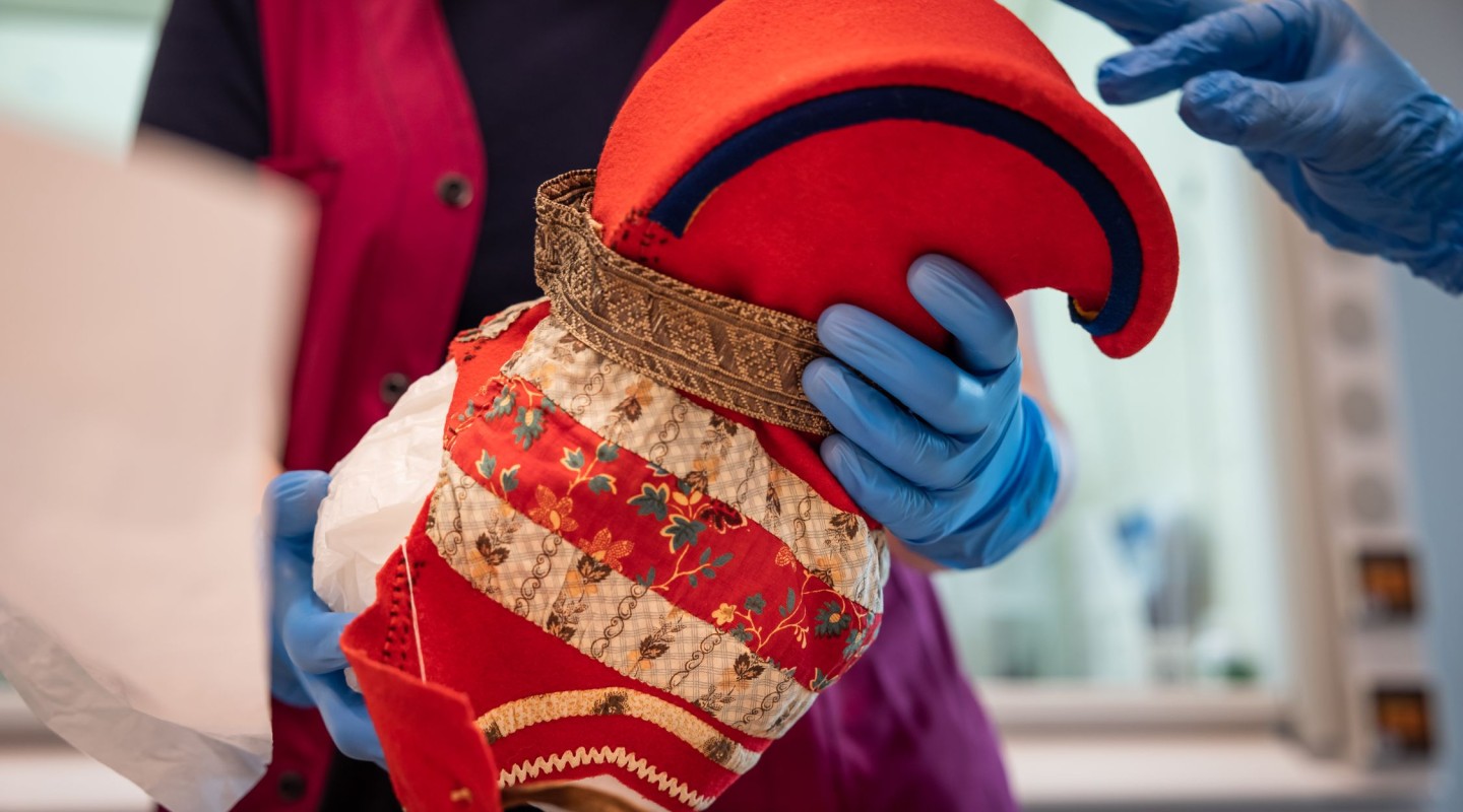 Hands wearing blue protective gloves are carefully handling a traditional "horn hat" with a red top and a decorative, patterned band. The person handling the hat is wearing a dark blue shirt and a red vest, suggesting they may be a professional conservator or curator in a museum setting. The focus on the gloves and the hat emphasizes the care being taken to preserve a culturally significant artifact.