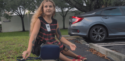 A person is seated on a curb in a parking lot, wearing a tie-dye dress and sandals, with a small blue handbag next to them. They have a tattoo on their arm and are wearing a name tag sticker. The person looks off to the side with a thoughtful expression. Behind them, there is a car parked and trees that suggest the area could be near an office or a public building. The setting appears to be during the day with overcast weather.