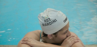 A swimmer is resting at the edge of a swimming pool, wearing a white swim cap with "R.O.T. MARDINI" printed on it alongside the Olympic rings and a logo for the brand "arena". The swimmer is leaning their arms on the poolside and has their face turned to one side, looking thoughtful or focused. Water droplets are visible on the swimmer's cap, indicating recent activity in the pool. The pool water has a bright blue color, which suggests an indoor setting with good lighting.