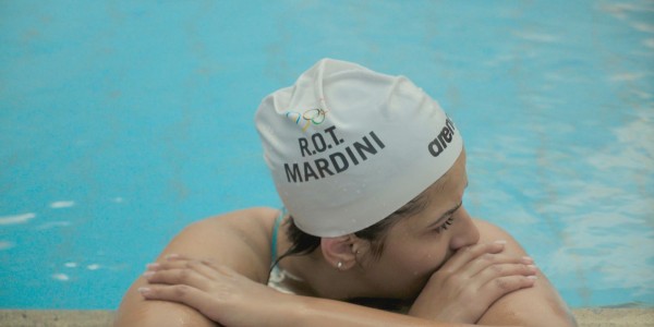 A swimmer is resting at the edge of a swimming pool, wearing a white swim cap with "R.O.T. MARDINI" printed on it alongside the Olympic rings and a logo for the brand "arena". The swimmer is leaning their arms on the poolside and has their face turned to one side, looking thoughtful or focused. Water droplets are visible on the swimmer's cap, indicating recent activity in the pool. The pool water has a bright blue color, which suggests an indoor setting with good lighting.