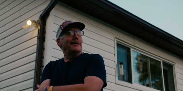 A person wearing glasses and a baseball cap is standing outside, arms crossed, with a slight smile on their face. They are looking off to the side, possibly at something out of the frame. The setting seems to be residential, as they are standing in front of a house with siding and a window reflecting the sky. It appears to be dusk, as the light is soft and a lamp next to the house is illuminated.