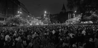 A dense crowd of people gathered at night on a city street, viewed from an elevated perspective. The black and white image captures the scene's high contrast, with streetlights illuminating the scene and a canopy tent visible in the distance. Buildings line the street and a spire looms in the background. The crowd's attention seems directed towards a point not visible in the image, possibly a stage or event out of frame.