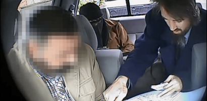 The interior of a vehicle with three individuals, captured seemingly from a dashboard camera. The face of the person in the front passenger seat is pixelated for privacy. In the back seat, one individual is leaning forward, looking at a map spread out over the center armrest, while another person in the back appears to be looking down, partially obscured from view. The individual examining the map has a beard and wears a dark jacket. The setting seems to be during the day, as daylight is visible through the car windows.