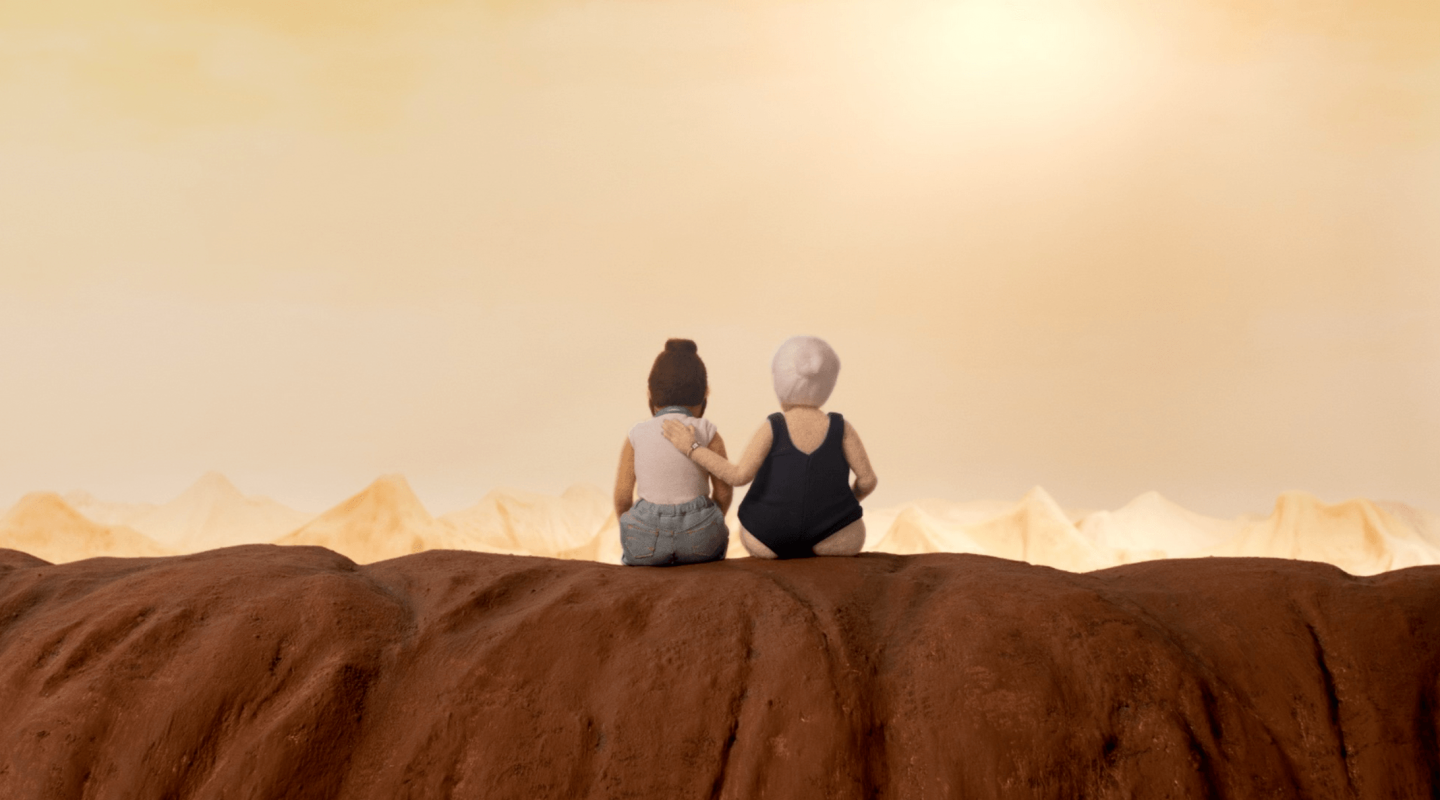 Two individuals are sitting close together on a high ledge, looking out over a desert-like landscape with sand dunes stretching towards the horizon. The sky is a warm, hazy orange, possibly indicating either sunrise or sunset. The companionship between the two figures is emphasized by one person's arm around the other, suggesting a moment of shared experience or reflection in a vast and tranquil environment.