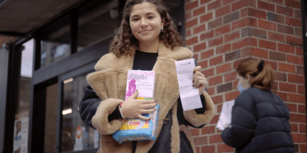 A woman stands outside a brick building, holding a box of Playtex tampons and a long printed receipt. They are smiling at the camera, dressed in a cozy, textured beige jacket with a navy blue top visible underneath. Their curly hair is pulled back from their face, and they wear round earrings.