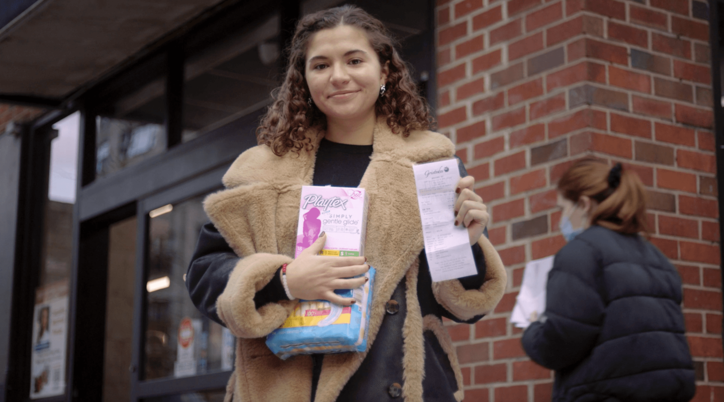 A woman stands outside a brick building, holding a box of Playtex tampons and a long printed receipt. They are smiling at the camera, dressed in a cozy, textured beige jacket with a navy blue top visible underneath. Their curly hair is pulled back from their face, and they wear round earrings.