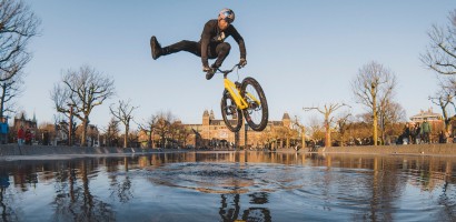 A BMX rider is captured mid-air performing a stunt, with their body horizontal to the ground and legs kicked out to the side, above a yellow bicycle. The bike's reflection is visible on the wet surface below. The shot is set against an urban park backdrop with historical buildings, bare trees, and a clear blue sky. Onlookers are seen in the distance. The low angle of the photograph emphasizes the height of the jump and the skill of the rider.