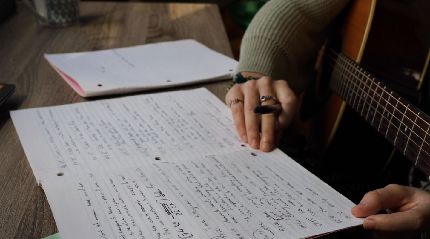 A close-up of someone's hands over a sheet of paper with handwritten notes, likely lyrics or music notes, as suggested by the presence of a guitar in the background. The person has several rings on their fingers and is holding a pen, possibly in the process of writing or editing the notes. The setting appears to be a wooden table, with another sheet of paper and a mug partially visible in the background, creating a casual and creative atmosphere.