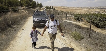 An adult and a child are walking hand in hand down a dusty rural path. The adult is wearing a white t-shirt with a logo, khaki pants, a cap, and sunglasses, and is carrying a backpack and a camera. The child is dressed in a striped school uniform and carrying a lollipop. In the background, there is a vehicle driving behind them. Beyond the vehicle stretches a landscape of rolling hills and sparse vegetation under a clear sky suggesting a warm, dry climate.