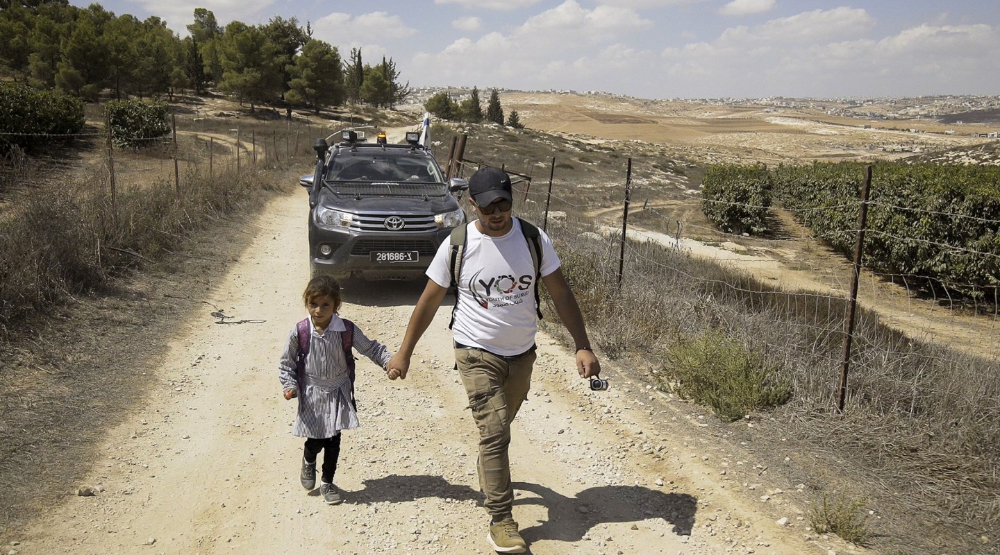 An adult and a child are walking hand in hand down a dusty rural path. The adult is wearing a white t-shirt with a logo, khaki pants, a cap, and sunglasses, and is carrying a backpack and a camera. The child is dressed in a striped school uniform and carrying a lollipop. In the background, there is a vehicle driving behind them. Beyond the vehicle stretches a landscape of rolling hills and sparse vegetation under a clear sky suggesting a warm, dry climate.