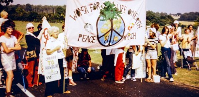 A group of people dressed in casual attire is gathered outdoors holding a banner that reads, "Women for Life on Earth Peace March '81". The banner features symbols of peace and the Earth. The setting is a sunny day with trees and open skies in the background.