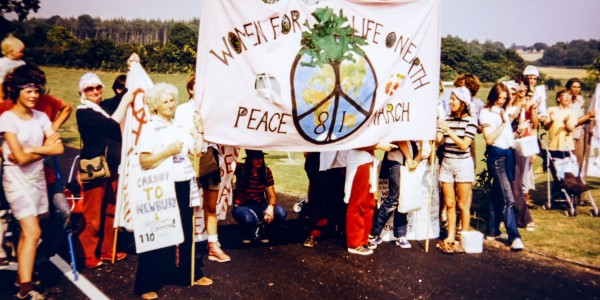 A group of people dressed in casual attire is gathered outdoors holding a banner that reads, "Women for Life on Earth Peace March '81". The banner features symbols of peace and the Earth. The setting is a sunny day with trees and open skies in the background.