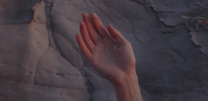 A hand with the palm facing forward and fingers slightly apart, set against a backdrop of a rock surface with natural lines and crevices. The lighting is soft and dim, suggesting either early morning or late afternoon, with warm tones that give the skin a reddish hue, contrasting with the cooler tones of the rock. The focus is on the hand, which is centered in the frame, while the rock surface in the background is slightly blurred. The composition is simple and evocative.
